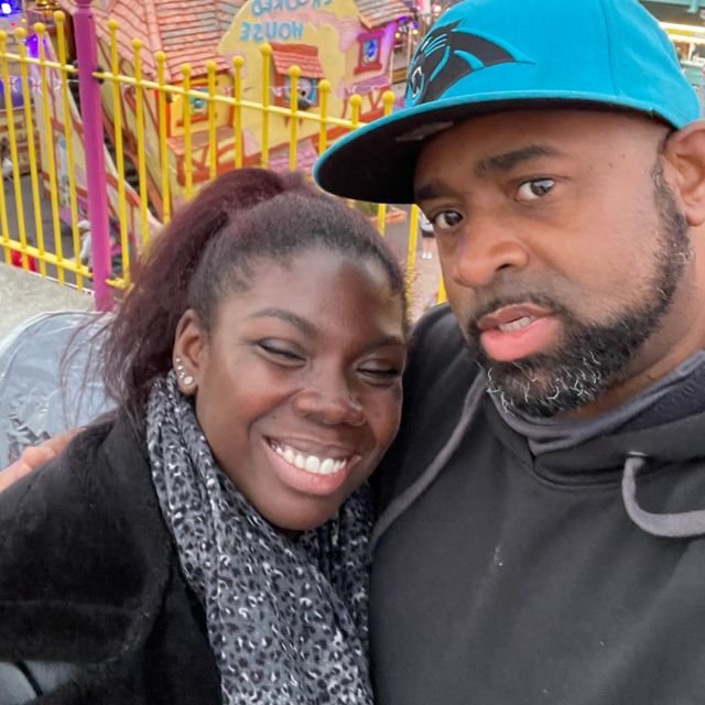 Bear and Carlene in a selfie at a seaside funfair, beside the Crooked House ride